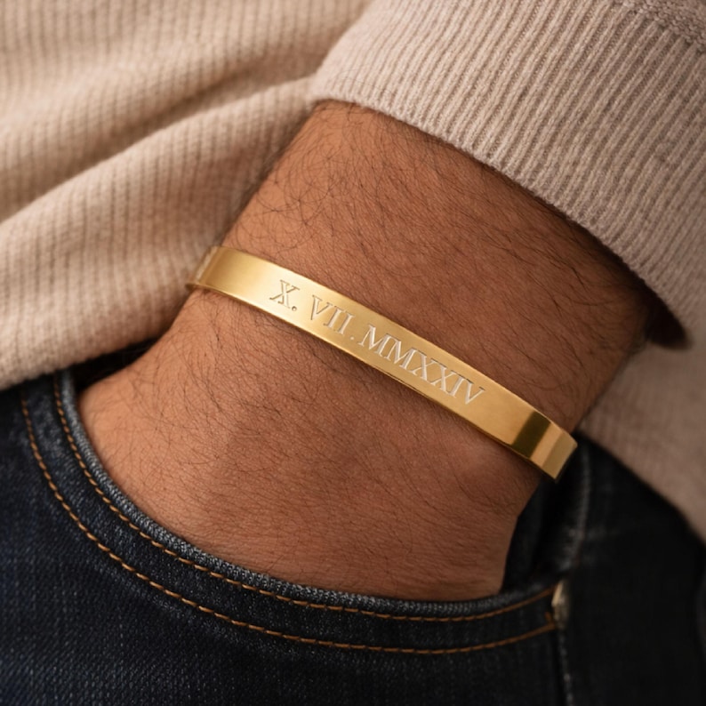 May include: A gold-toned metal cuff bracelet featuring a smooth, polished finish. The outer surface of the bracelet is engraved with the Roman numerals X. VII. MMXXIV in a clean, sans-serif font, centred along the length of the band.