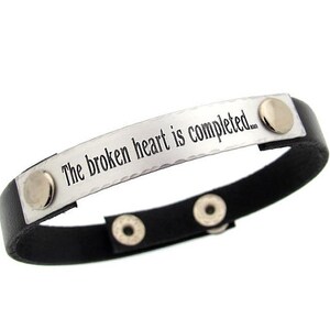 May include: A black leather bracelet with a silver metal plate engraved with the words "The broken heart is completed..."