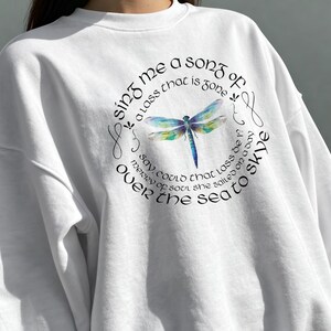 Puede incluir: Sudadera blanca con un diseño de libélula en acuarela dentro de una disposición de texto circular. El texto dice: "Sing me a song of a lass that is gone over the sea to Skye."