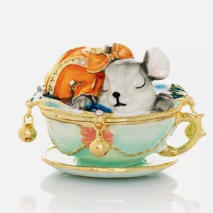 Könnte beinhalten: Eine kleine, weiße Mausfigur schläft in einer Teetasse mit Untertasse. Die Teetasse ist mit einem Blumendesign und goldenen Verzierungen verziert. Die Maus trägt einen kleinen, orangefarbenen und goldenen Hut.