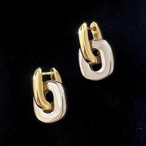 Puede incluir: Un par de pendientes de eslabones de cadena dorados y plateados. Los pendientes tienen un cierre de color dorado y un diseño de eslabón de cadena plateado. Los pendientes se muestran sobre un fondo negro.