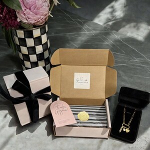 May include: An open cardboard box with a gold wax seal and a pink gift box with a black ribbon. A gold necklace with heart charms is displayed in a black velvet box. A vase with pink flowers is in the background.