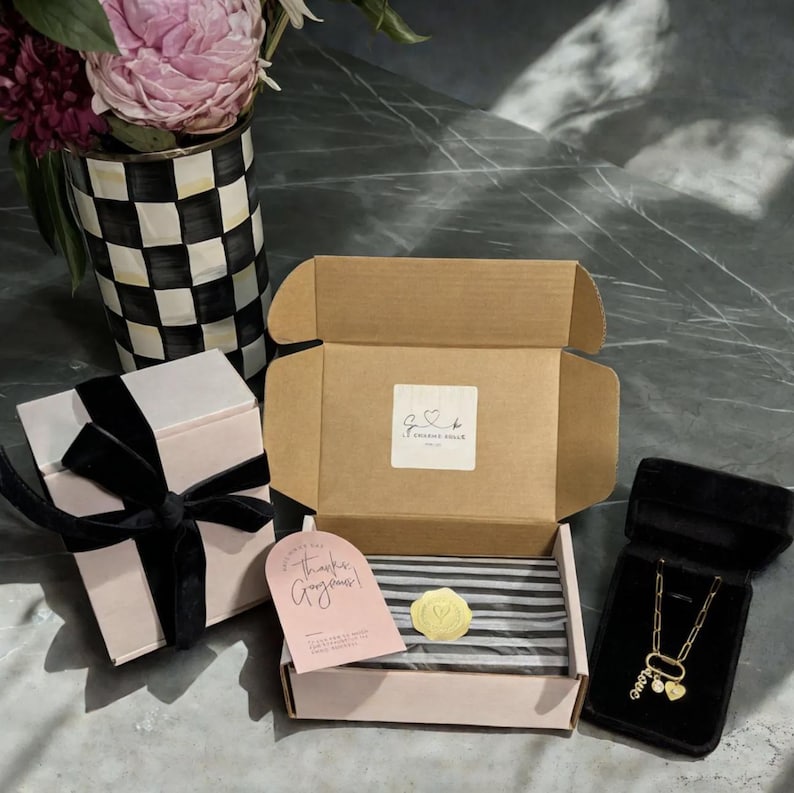 May include: An open cardboard box with a gold wax seal and a pink gift box with a black ribbon. A gold necklace with heart charms is displayed in a black velvet box. A vase with pink flowers is in the background.