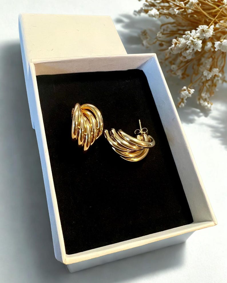 Puede incluir: Un par de pendientes dorados presentados en una caja de joyer&iacute;a crema abierta con interior negro. Los pendientes tienen un dise&ntilde;o de m&uacute;ltiples aros. La caja est&aacute; sobre una superficie blanca, con un arreglo floral borroso en el fondo.