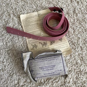 Op de afbeelding: Een opgerolde, stoffig roze rubberen tourniquet met een reeks gaten en een zwarte sluiting. Daarnaast ligt een gevouwen papier met tekst en illustraties, samen met een verzegeld, grijs pakket met Cyrillische tekst.