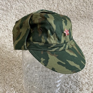 Puede incluir: Un gorro de camuflaje de estilo militar con orejeras y un emblema de estrella roja. El gorro presenta un patrón verde, marrón y beige. El gorro está hecho de una tela duradera y tiene un diseño estructurado. Es un accesorio de estilo militar.