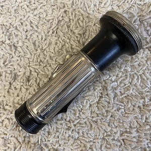 May include: A vintage flashlight with a black body and a silver-coloured, ribbed metal casing. The flashlight has a textured end and a small clip on the side. The top of the flashlight has a silver-coloured, ridged bezel.