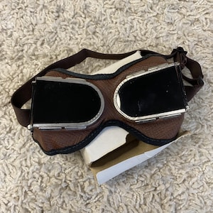 Könnte beinhalten: Braune Leder-Fliegerbrille mit schwarzen Gläsern und einem braunen Riemen. Die Brille hat einen silbernen Metallrahmen um die Gläser und eine braune Leder-Gesichtsmaske. Die Brille liegt auf einer weißen und braunen Pappschachtel.