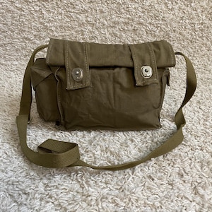 May include: An olive green canvas shoulder bag with a flap closure and two metal snaps. It features a long, adjustable strap and a small side pocket. The bag is designed for carrying items.