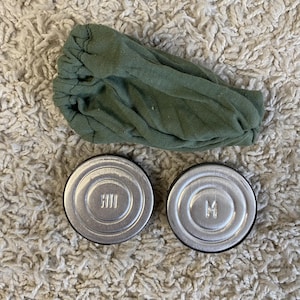 May include: Two silver-colored, round containers with black rims and embossed letters, next to a green fabric item. The containers have the letters "HH" and "M" embossed on their lids. The green fabric is gathered and appears to be a headband or similar accessory.