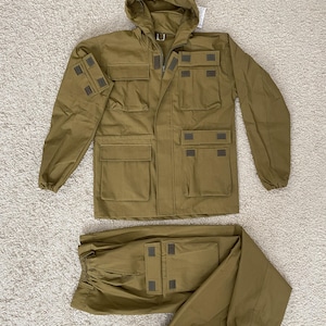 Può includere: Un completo giacca e pantaloni in stile militare color kaki. La giacca ha un cappuccio, tasche multiple e toppe. I pantaloni hanno una vita elasticizzata e polsini. Il completo è realizzato in tessuto resistente.