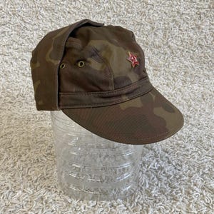 Puede incluir: Gorra con estampado de camuflaje con orejeras y un emblema de estrella roja. El sombrero presenta un diseño de camuflaje marrón, verde y beige. La estrella roja está en la parte delantera del sombrero. El sombrero está sobre una botella de plástico transparente.