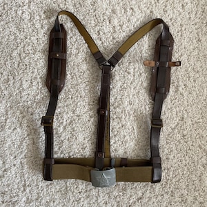 May include: Brown leather and olive green canvas military suspenders with a silver-colored buckle featuring a star. The suspenders have adjustable straps and shoulder pads. The design includes a belt and a Y-shaped back.