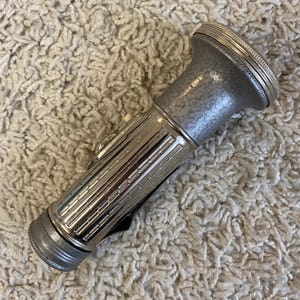 May include: A vintage silver flashlight with a textured body and a ribbed design. The flashlight has a push button and a clip on the side. The top and bottom of the flashlight have a circular design.