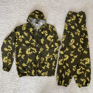 Peut inclure: Un ensemble de camouflage en deux pièces, comprenant une veste à capuche avec une fermeture éclair et un pantalon assorti. L'ensemble est orné de formes pixélisées vert olive et jaune-beige. La veste a une capuche à cordon et le pantalon une taille élastique.