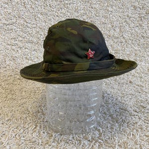 Puede incluir: Sombrero boonie con estampado de camuflaje y un emblema de estrella roja. El sombrero tiene un ala ancha y una correa para la barbilla. El sombrero está hecho de una tela duradera y está diseñado para uso en exteriores. El sombrero es de estilo militar.