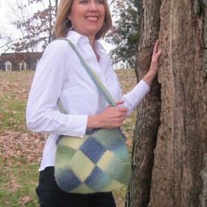 May include: A woman wearing a white button-down shirt with a blue and green crocheted bag slung over her shoulder. She is standing next to a tree trunk.
