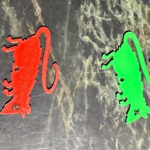 May include: Two rat-shaped ornaments, one red and one green, are displayed against a mottled gray background. Each rat has a long tail, small legs, and a star-shaped detail. The ornaments appear to be 3D printed.