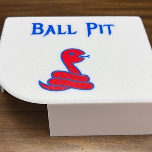 May include: A white plastic cover featuring the text BALL PIT in blue lettering above a red snake graphic with a blue outline. The object has a flat top surface with one rounded corner and a vertical side panel extending downward from the edge.
