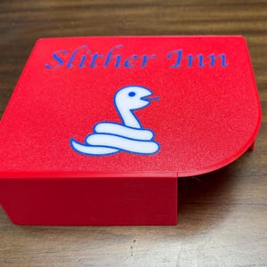 May include: A bright red, flat-topped plastic object with a curved corner, featuring the blue text Slither Inn and a white graphic of a coiled snake with a blue outline printed on the top surface, designed with a vertical side panel.