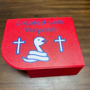 May include: A red rectangular box with a rounded corner featuring the blue text Created with Purpose. The top surface displays a white snake graphic coiled in the center, flanked by two blue cross symbols, one on each side of the snake illustration.