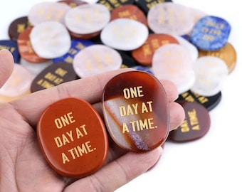 May include: Two red agate stones with gold lettering. The stones are smooth and oval shaped. The text on the stones reads "One Day At A Time."