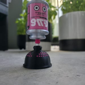 Könnte beinhalten: Ein zylindrisches, rosa-silbernes Objekt mit einem Cartoon-Gesicht und dem Text "S11" auf der Seite, das vertikal auf einer schwarzen, kuppelförmigen Basis mit perforierter Oberfläche montiert ist. Das Objekt hat ein kleines rosa Verbindungsstück.