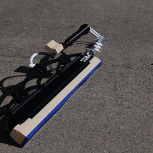 May include: A black and beige squeegee featuring a blue rubber blade, a black handle, and a white coiled spring mechanism. The word "KITZ" is printed in white on the black frame. The tool includes a black plastic structure with a beige block.