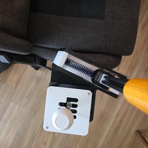 May include: A black and white device with a white knob and a gear shift pattern. The device is attached to a black and yellow arm, and is positioned near a gray upholstered chair. The device appears to be a controller.