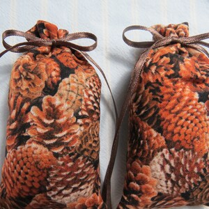 May include: Two small fabric pouches with a repeating pattern of brown and orange feathers. Each pouch is tied with a brown ribbon, forming a bow at the top. The pouches are likely used for storing small items or as sachets.