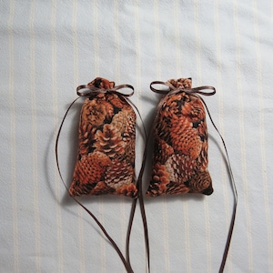 May include: Two small, fabric sachets with a brown pine cone pattern. Each pouch is tied with a brown ribbon and has a long, thin ribbon hanging down. The sachets are on a light blue and white striped background.