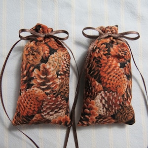 May include: Two small, rectangular fabric sachets with a brown pine cone pattern. Each sachet is tied with a brown ribbon. The background is a light blue and white striped fabric.