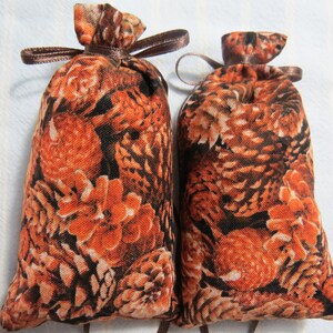 May include: Two small, fabric pouches with a repeating pattern of orange and brown pine cones and foliage. Each pouch is tied at the top with a brown ribbon. The pouches are likely filled with potpourri or other scented items.