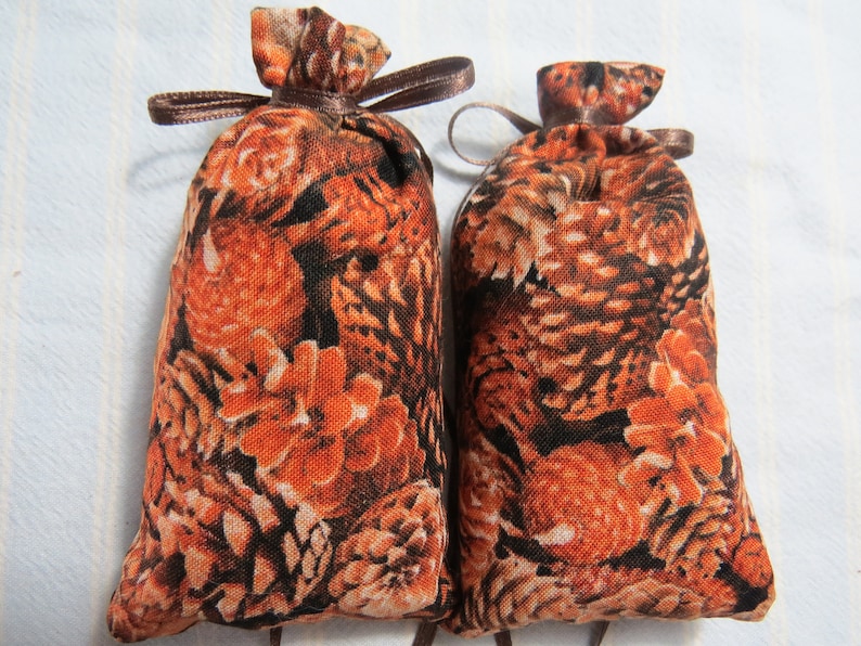 May include: Two small, fabric pouches with a repeating pattern of orange and brown pine cones and foliage. Each pouch is tied at the top with a brown ribbon. The pouches are likely filled with potpourri or other scented items.