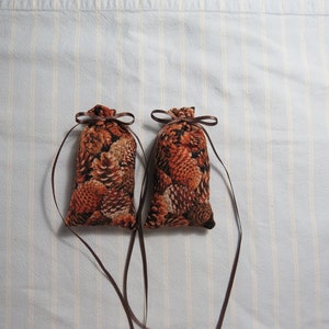 May include: Two small, fabric sachets with a brown pine cone pattern. Each sachet is tied with a brown ribbon and has a matching ribbon extending from the top. The background is a light blue and white striped fabric.
