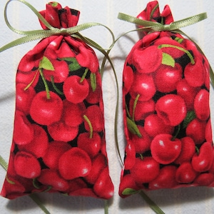 以下が含まれることがあります： 緑のリボンで結ばれた赤い布製のサシェが2つ。サシェはチェリー柄の布で満たされています。