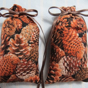 May include: Two small fabric pouches with a brown and orange pinecone pattern. Each pouch is tied at the top with a brown ribbon, forming a bow. The pouches are likely used for storage or as decorative items.