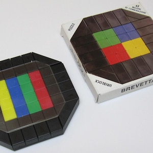 Könnte beinhalten: Ein schwarzes und braunes Holzpuzzle mit einer durchsichtigen Plastikabdeckung. Das Puzzle hat die Form eines Achtecks und ein quadratisches Design in der Mitte. Das Quadrat besteht aus vier kleineren Quadraten, jedes in einer anderen Farbe: rot, gelb, grün und blau. Das Puzzle ist mit "Uriblock" und "Brevettato" beschriftet.