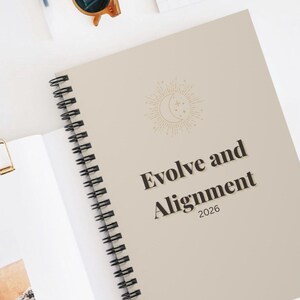May include: A spiral-bound planner with a beige cover featuring the text "Evolve and Alignment 2026". The cover also includes a celestial design with a crescent moon and stars. The planner is open to reveal blank pages.