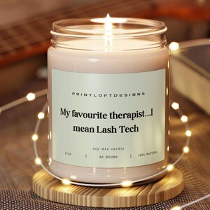 May include: A lit soy wax candle in a glass jar with a label that reads "My favourite therapist...I mean Lash Tech." The candle is on a wooden coaster and surrounded by string lights. The candle is 255 grams and burns for 60 hours.
