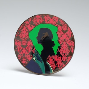 May include: Round decorative pin with a black silhouette of a person's profile, set against a green backdrop. The pin features a red and black damask pattern on the outer ring. The design includes a figure within the profile.