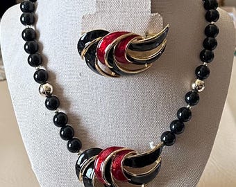 Puede incluir: Un conjunto de joyas vintage con un collar de cuentas negras con detalles plateados y un broche a juego. El broche y el colgante del collar tienen un diseño de metal dorado con detalles de esmalte negro y rojo. El conjunto se exhibe en un maniquí.