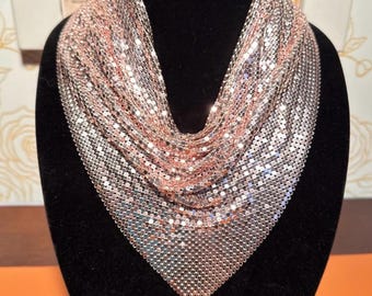 May include: A rose gold-toned mesh scarf, displayed on a black mannequin head. The scarf is composed of small, interconnected metallic discs, creating a fluid, draped effect. The scarf's design is triangular, suitable for formal or casual wear.