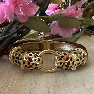 Op de afbeelding: Gouden scharnierende armband met luipaardenkopaccenten. De luipaardenkoppen hebben zwarte vlekken en rode ogen. De armband heeft een bruine emaille band en een gouden sluiting. Roze bloemen staan op de achtergrond.
