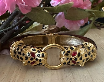 Op de afbeelding: Gouden scharnierende armband met luipaardenkopaccenten. De luipaardenkoppen hebben zwarte vlekken en rode ogen. De armband heeft een bruine emaille band en een gouden sluiting. Roze bloemen staan op de achtergrond.