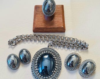 May include: A silver-toned jewellery set comprising a ring, necklace, and two pairs of earrings. Each piece features an oval, dark-coloured stone set within a decorative silver frame. The ring is displayed on a wooden stand.