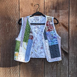Op de afbeelding: Een patchwork vest met een witte achtergrond en kleurrijke bloemen- en geometrische patronen. Het vest is gemaakt van stofresten die in een willekeurig patroon aan elkaar zijn genaaid. Het hangt aan een houten hanger tegen een houten achtergrond.