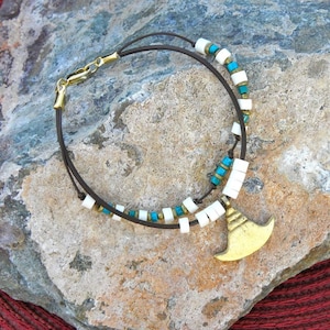 May include: A handcrafted bracelet featuring a brown leather cord, adorned with white and turquoise beads, and gold-colored accents. The bracelet includes a gold-colored charm and a gold-colored clasp.