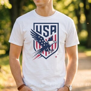 May include: White t-shirt featuring a shield design with the word "USA" at the top. The shield showcases a soaring eagle with stars and stripes in red, white, and blue. A patriotic design for fans of the USA.
