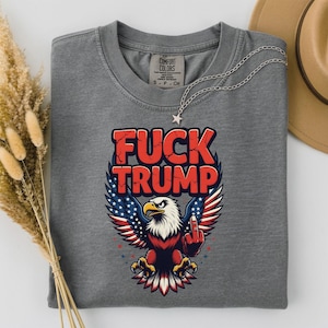 Op de afbeelding: Grijs t-shirt met de tekst "FUCK TRUMP" in het rood, boven een adelaarsafbeelding met vleugels in de kleuren van de Amerikaanse vlag. De adelaar steekt zijn middelvinger op. Een zilveren ketting met een sterhanger ligt op het shirt.